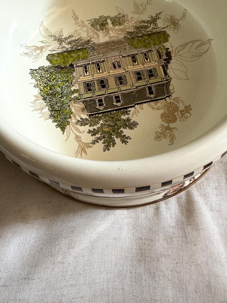 May include: A cream-colored, vintage-style bowl with a detailed illustration of a house surrounded by trees. The bowl's rim features a checkered pattern with gold accents, and floral designs. The overall aesthetic is reminiscent of antique tableware.