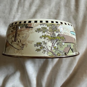 May include: A vintage metal cake dome with a cream-colored exterior. The dome features a detailed pastoral scene with trees, houses, and figures in shades of brown, green, and blue. The rim has a gold-colored trim and a series of black squares.