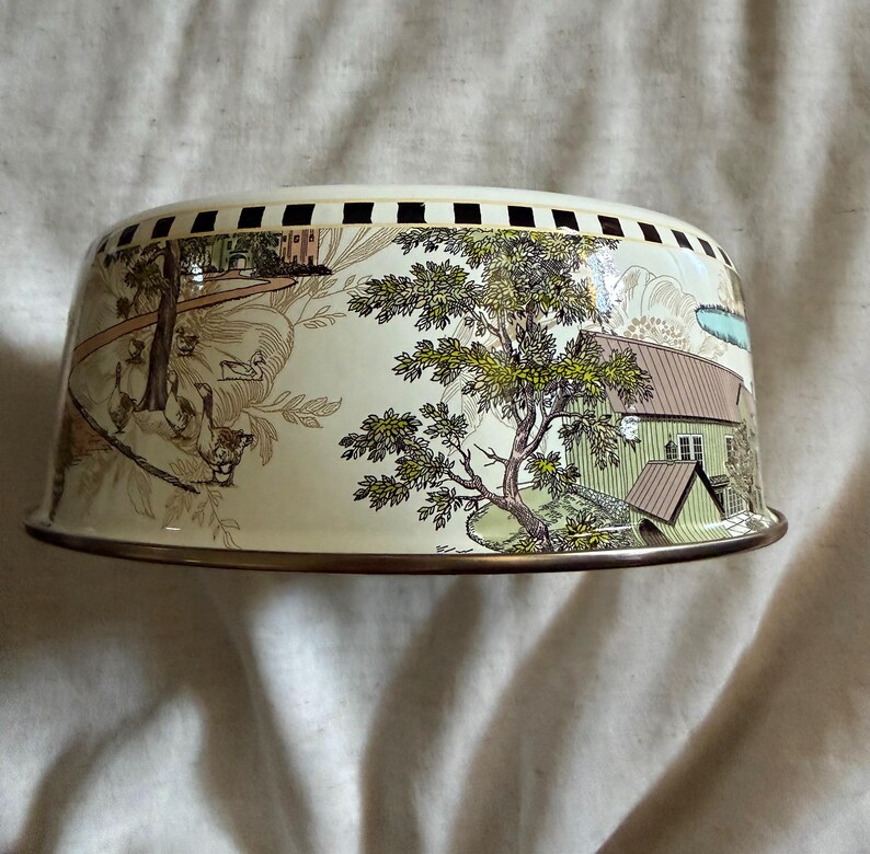 May include: A vintage metal cake dome with a cream-colored exterior. The dome features a detailed pastoral scene with trees, houses, and figures in shades of brown, green, and blue. The rim has a gold-colored trim and a series of black squares.