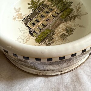 May include: A cream-colored ceramic bowl with a detailed illustration of a house and foliage. The bowl's interior features a house with green shutters and a dark roof, surrounded by green trees and floral accents. The rim has a black and white checkered pattern.