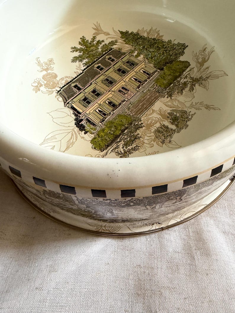 May include: A cream-colored ceramic bowl with a detailed illustration of a house and foliage. The bowl's interior features a house with green shutters and a dark roof, surrounded by green trees and floral accents. The rim has a black and white checkered pattern.