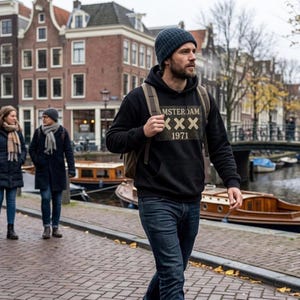 Peut inclure: Un sweat à capuche noir avec le texte "MSTERDAM XXX 1971" dans un design rectangulaire. La personne sur l'image porte un bonnet bleu, un jean bleu foncé et un sac à dos. L'arrière-plan montre un canal et des bâtiments.