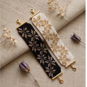 May include: Two rectangular beaded bracelets featuring a repeating floral pattern. One bracelet is black with gold beads, and the other is white with gold beads. Both bracelets have gold-toned metal end bars and lobster claw clasps at each end.