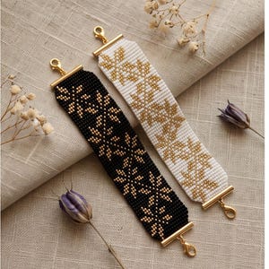 Può includere: Due bracciali larghi di perline con un motivo floreale ripetuto. Un bracciale è realizzato con perline nere e accenti dorati, l'altro con perline bianche e accenti dorati. Entrambi i bracciali presentano terminali in metallo dorato e moschettoni.