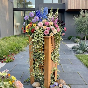 May include: A wooden planter box with a lattice design, overflowing with colorful flowers and trailing ivy. The arrangement includes roses, delphiniums, and other blooms in shades of pink, purple, yellow, and red. Decorative stones are at the base.