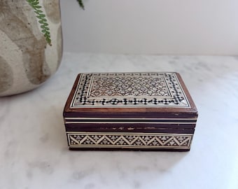 Könnte beinhalten: Eine rechteckige Holzschatulle mit aufwendigen geometrischen Einlegearbeiten auf dem Deckel und der Vorderseite. Die dunkle Holzoberfläche ist mit hellen, perlmuttartigen dreieckigen und rautenförmigen Mustern in einem sich wiederholenden Mosaikdesign verziert.