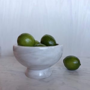 May include: A white marble pedestal bowl with a smooth, rounded shape and a flared base. Several bright green limes are nestled inside the bowl, and one additional lime rests on the surface next to the base of the marble vessel.