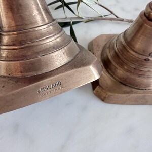 K&ouml;nnte beinhalten: Zwei Messingkerzenst&auml;nder mit konischer Form und gestuftem Sockel. Auf dem Sockel eines Kerzenst&auml;nders ist der Text ENGLAND sowie eine Zahlenreihe eingepr&auml;gt. Das Metall hat eine warme, gealterte Patina mit sichtbarer Oberfl&auml;chenstruktur.