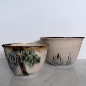 May include: Two ceramic bowls with a light beige base and a dark brown rim. The smaller bowl features a painted green tree design, while the larger bowl displays a painted cluster of brown cattails and green foliage on the exterior surface.