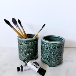 May include: Two cylindrical dark green ceramic brush holders with embossed relief patterns of landscapes and figures. One holder contains four wooden-handled paint brushes with dark bristles. Two tubes of Daniel Smith watercolor paint lie in front.