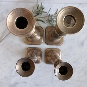 K&ouml;nnte beinhalten: Ein Set aus vier Messingkerzenhaltern, die in einer quadratischen Formation angeordnet und von oben fotografiert sind. Jeder Kerzenhalter hat eine kreisf&ouml;rmige &Ouml;ffnung, einen gerippten zylindrischen Schaft und einen quadratischen Fu&szlig;.