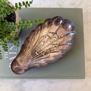 May include: A silver-toned metal dish shaped like a seashell with a fluted, scalloped edge. The dish contains several gold-colored rings and a beaded bracelet. The surface of the metal shows a tarnished, aged patina with dark and light highlights.
