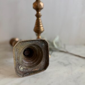 K&ouml;nnte beinhalten: Ein Messing-Kerzenhalter mit quadratischem Fu&szlig;, einer vertieften Mitte und einem hohen, schlanken Schaft mit dekorativen gedrechselten Details. Die Metalloberfl&auml;che weist eine verwitterte, gealterte Patina mit sichtbaren Oxidationsspuren auf.