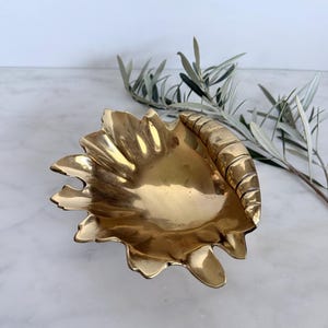 May include: A polished brass bowl shaped like a seashell with a scalloped edge and a ribbed, curved handle section. The metallic surface reflects light, highlighting the organic, textured form of the shell design in a shiny gold tone.