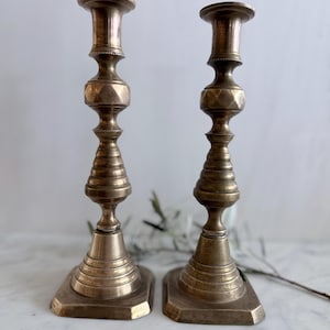 K&ouml;nnte beinhalten: Ein Paar hohe, metallische Messingkerzenst&auml;nder mit einem mehrstufigen, gedrehten Design, einem facettierten sechseckigen Mittelteil und einem breiten, quadratischen Fu&szlig;. Die Oberfl&auml;che hat ein mattes, gealtertes Finish mit sichtbaren Rillen.
