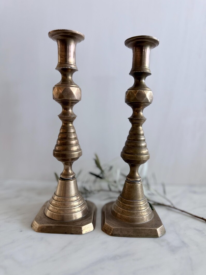 K&ouml;nnte beinhalten: Ein Paar hohe, metallische Messingkerzenst&auml;nder mit einem mehrstufigen, gedrehten Design, einem facettierten sechseckigen Mittelteil und einem breiten, quadratischen Fu&szlig;. Die Oberfl&auml;che hat ein mattes, gealtertes Finish mit sichtbaren Rillen.
