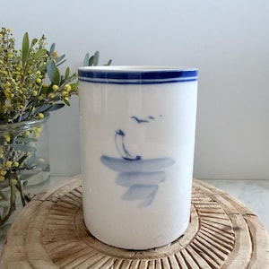 May include: A cylindrical white ceramic vessel featuring a hand-painted blue sailboat motif on the front and a horizontal blue band around the top rim. The design depicts a simple blue boat on water with a small bird silhouette above it.