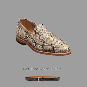 Pode incluir: Um sapato estilo mocassim com padrão de pele de cobra em tons de bege, castanho e preto. O sapato tem bico arredondado e sola castanha. Uma correia de couro castanho com fivela prateada também é mostrada.