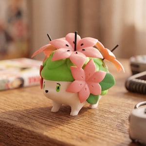 May include: A small, white, four-legged creature figurine with a green, leafy back adorned with several large, light pink, star-shaped flowers. The figure has a smooth, matte finish, a small black nose, and bright green eyes, standing in a neutral pose.