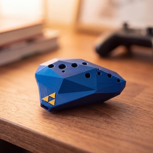 May include: A blue ocarina with a geometric, faceted surface and multiple finger holes. The instrument features a small, yellow triangular emblem on its front side, consisting of three smaller triangles arranged in a larger triangular pattern.