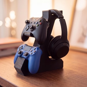 May include: A black 3D-printed gaming stand designed to hold two controllers and a pair of over-ear headphones. The stand features a top cradle for a black Xbox controller, a lower cradle for a blue PlayStation controller, and a side hook for headphones.