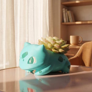 May include: Three character-themed planters on a wooden surface. One is a light green creature in a brown terracotta pot with a single leaf. The second is a blue turtle-like figure. The third is a teal creature with a plant growing from its back.