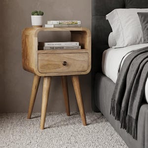 Peut inclure: Une table de chevet en bois clair aux bords arrondis, dotée d'une étagère de rangement ouverte et d'un tiroir unique avec un petit bouton rond. La table repose sur quatre pieds fuselés et supporte un petit cactus en pot et deux livres.
