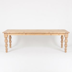 May include: A rectangular dining table crafted from light-toned natural wood. The table features a smooth, flat top surface with visible wood grain patterns and is supported by four sturdy legs, each designed with a decorative turned spindle shape.
