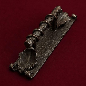 Pode incluir: Um puxador de porta de metal escuro com uma pega texturizada em forma de ramo envolvida por anéis metálicos. O puxador está montado numa placa retangular com acabamento martelado, decorada com duas folhas de metal esculpidas nas extremidades.