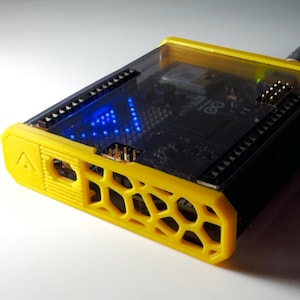 Op de afbeelding: Een zwarte Arduino-microcontrollerkaart in een felgele, 3D-geprinte beschermende behuizing met een honingraatpatroon aan de zijkant en een doorschijnende bovenklep, met een klein blauw LED-lampje dat door de behuizing heen zichtbaar is.