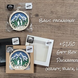 May include: A wooden ornament featuring a mountain scene with two hikers and the word "Colorado" in a circle. The ornament is shown in three different packaging options: basic, kraft gift box, and black or white gift box. The text "+$2.50 Gift Box Packaging (Kraft, Black, White)" is also shown.
