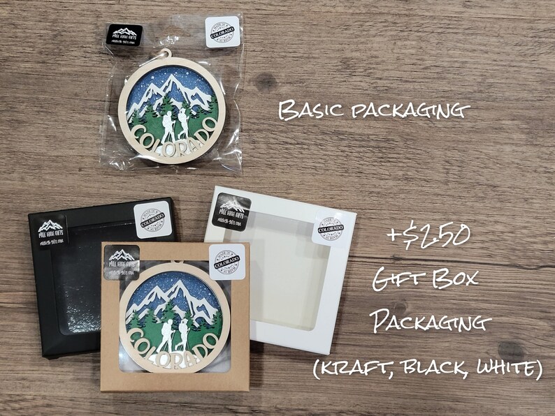 May include: A wooden ornament featuring a mountain scene with two hikers and the word "Colorado" in a circle. The ornament is shown in three different packaging options: basic, kraft gift box, and black or white gift box. The text "+$2.50 Gift Box Packaging (Kraft, Black, White)" is also shown.