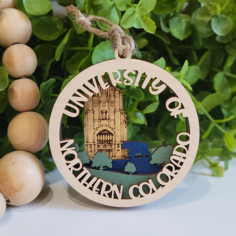 May include: A wooden ornament with a cutout design featuring the University of Northern Colorado logo. The logo includes the university's name, a building, and a bear.