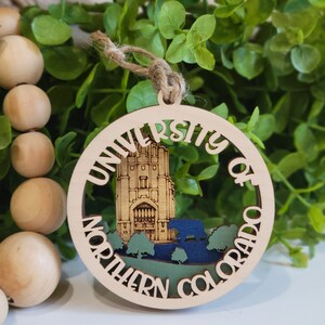 May include: A wooden ornament with a cutout design of the University of Northern Colorado campus. The ornament is painted in shades of brown, green, and blue. The text "University of Northern Colorado" is written around the edge of the ornament.