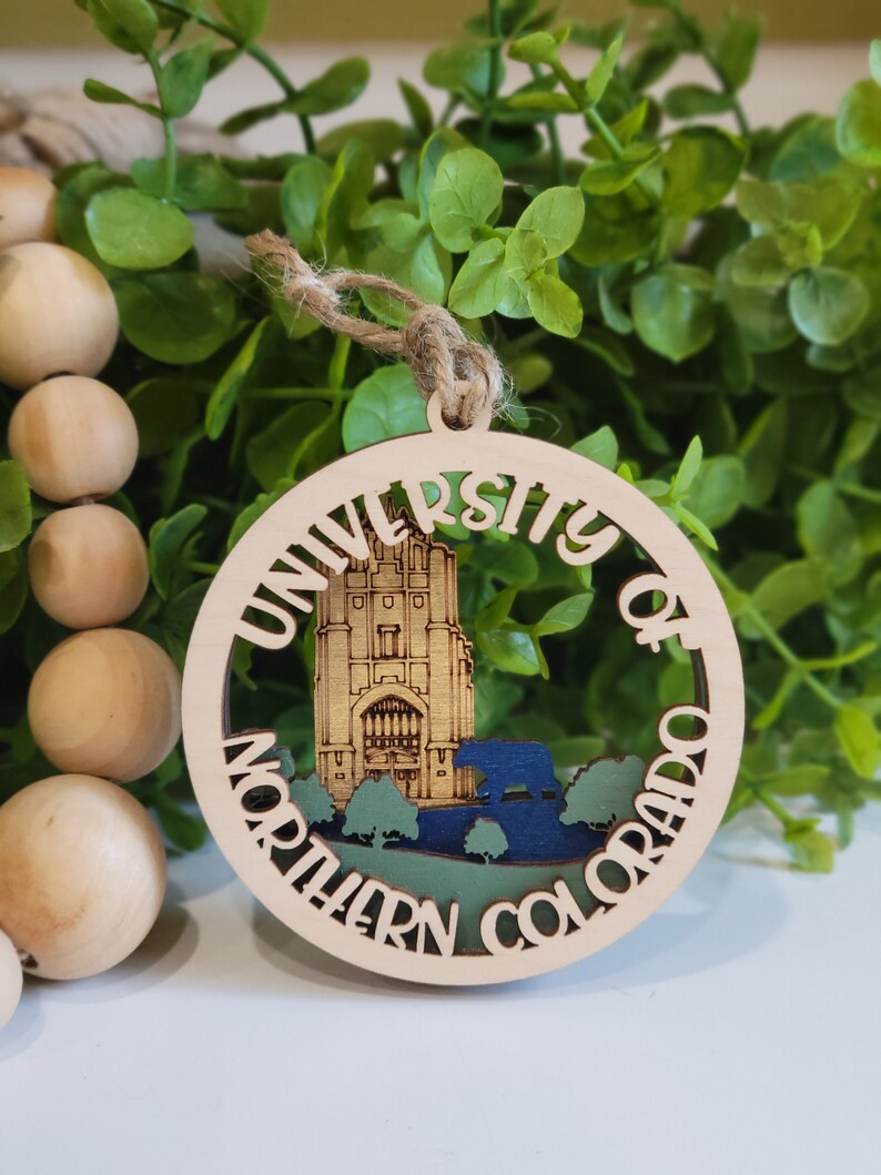 May include: A wooden ornament with a cutout design of the University of Northern Colorado campus. The ornament is painted in shades of brown, green, and blue. The text "University of Northern Colorado" is written around the edge of the ornament.