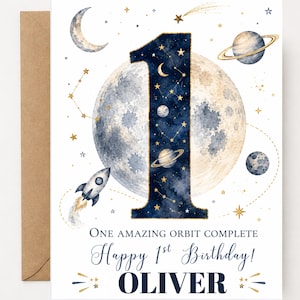 May include: A white greeting card featuring a large number one filled with a dark blue starry night sky pattern. The design includes a moon, planets, a rocket, and gold stars. Text reads: ONE AMAZING ORBIT COMPLETE Happy 1st Birthday! OLIVER.