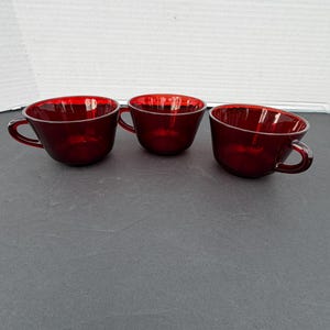 May include: Three matching ruby red glass teacups arranged in a horizontal row. Each cup features a smooth, translucent finish, a rounded bowl shape, and a small, curved handle attached to the side for holding. The glass is a deep, vibrant red.