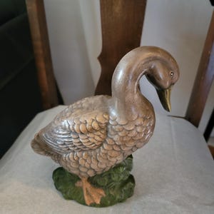 May include: A decorative figurine of a duck with a brown, textured body and detailed feather patterns. The duck has a smooth, curved neck, a pointed beak, and is standing on a green, textured base that resembles grass or foliage.