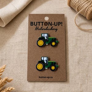 May include: Two green and yellow tractor-shaped buttons on a brown card. The card reads "BUTTON-UP! Haberdashery" and "button-up.ca". The buttons are detailed with black outlines and white windows. A spool of twine and fabric are in the background.