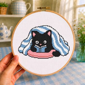 May include: A circular wooden embroidery hoop containing a cross-stitch design of a black cat with white eyes and pink ears, tucked under a blue and white striped blanket while resting on a pink cushion against a white fabric background.