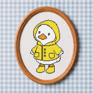 May include: An oval wooden frame containing a cross-stitch embroidery of a white duckling wearing a yellow hooded raincoat and matching yellow rain boots. The duckling has an orange beak and is depicted against a white fabric background.