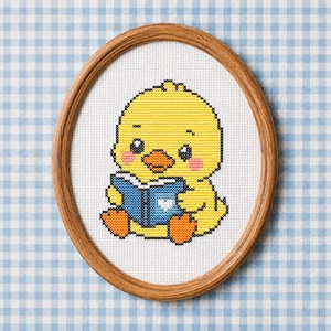 Può includere: Un ricamo a punto croce raffigurante un pulcino giallo con zampe e becco arancioni, che tiene in mano un libro blu con un dettaglio a forma di cuore bianco. Il disegno è ricamato su tessuto bianco e racchiuso in una cornice ovale in legno.