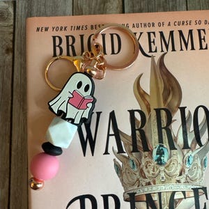May include: A rose gold keychain with a ghost charm reading a book. The keychain has a pink, black, and white beaded accent. The keychain is next to a book with the title "Warrior Princess".
