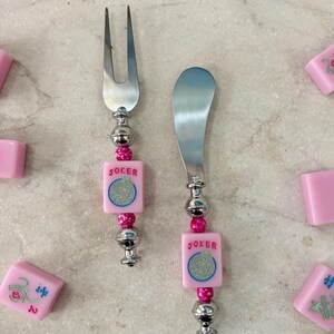 May include: A cheese fork and spreader set with pink and silver accents. The handles feature pink square tiles with the word "JOKER" and a circular design. The set is surrounded by pink tiles.