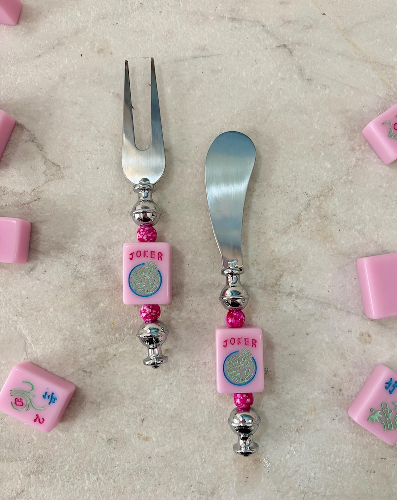 May include: A cheese fork and spreader set with pink and silver accents. The handles feature pink square tiles with the word "JOKER" and a circular design. The set is surrounded by pink tiles.