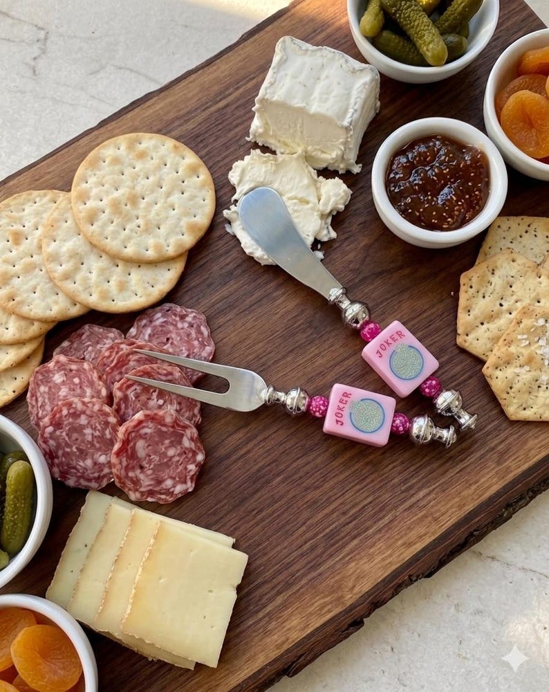May include: A wooden charcuterie board with an assortment of snacks, including crackers, cheese, salami, and pickles. Two serving utensils with pink "Joker" accents are also present. The board is ideal for entertaining.