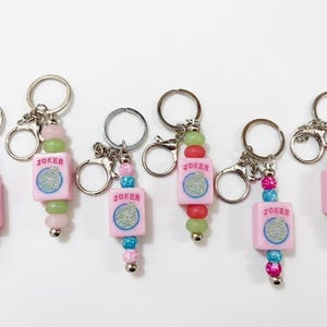 May include: Keychains with pink square charms that read "JOKER" with a glittery design. The charms are strung with colorful beads in shades of pink, green, red, and blue. Each keychain has a silver keyring and clasp.