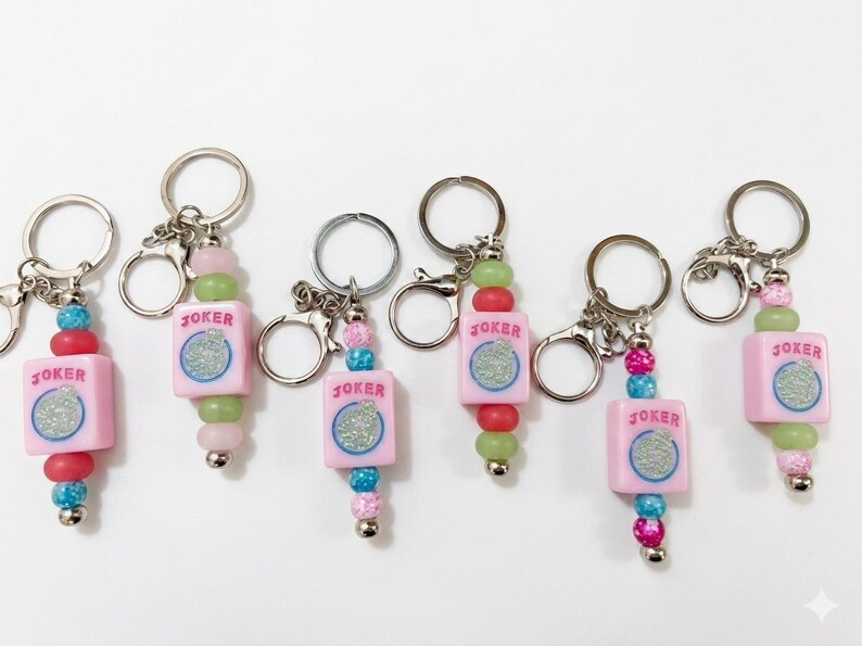 May include: Keychains with pink square charms that read "JOKER" with a glittery design. The charms are strung with colorful beads in shades of pink, green, red, and blue. Each keychain has a silver keyring and clasp.