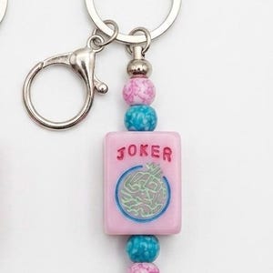 May include: Pink and blue beaded keychain with a silver clasp. The keychain features a pink rectangular charm with the word "JOKER" and a circular design. The beads are pink, blue, and silver.