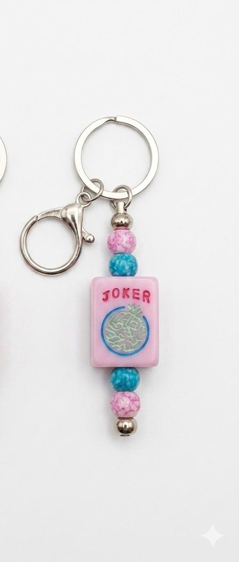 May include: Pink and blue beaded keychain with a silver clasp. The keychain features a pink rectangular charm with the word "JOKER" and a circular design. The beads are pink, blue, and silver.
